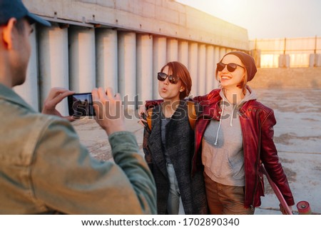 Man taking pictures of two young happy grimacing women with short hair in sunglasses with skateboards. Sunset in a city. Gray floor and building.