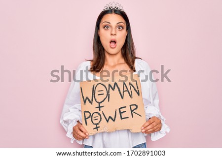 Girl wearing princess tiara asking for women rights holding banner with woman power message scared and amazed with open mouth for surprise, disbelief face