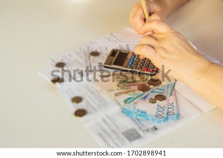 an elderly woman considers payments for utility bills; on a calculator, she considers payment for an apartment with money in Russian rubles; banknotes of 2000,100 rubles and Russian coins Royalty-Free Stock Photo #1702898941