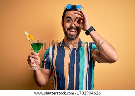Young handsome tourist man with beard on vacation wearing summer shirt drinking cocktail with happy face smiling doing ok sign with hand on eye looking through fingers