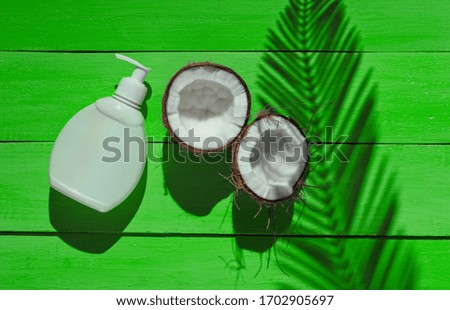 Minimalistic beauty still life. Two halves of chopped coconut and white bottle of cream with shadows from palm leaves on green wooden background. Creative fashion concept. Top view.