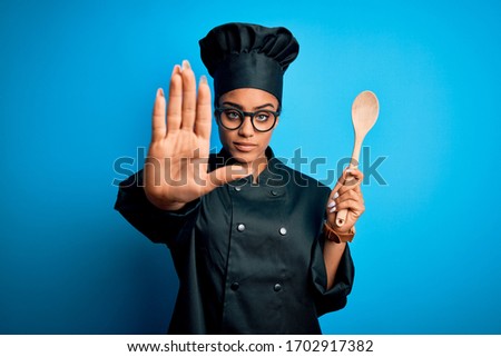 Young african american chef girl wearing cooker uniform and hat holding wooden spoon tool with open hand doing stop sign with serious and confident expression, defense gesture