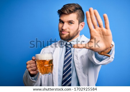 Young blond doctor man with beard and blue eyes wearing coat drinking jar of beer with open hand doing stop sign with serious and confident expression, defense gesture