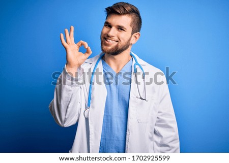 Young blond doctor man with beard and blue eyes wearing white coat and stethoscope smiling positive doing ok sign with hand and fingers. Successful expression.