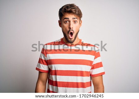 Young handsome man with beard wearing striped t-shirt standing over white background afraid and shocked with surprise expression, fear and excited face.