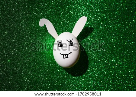 White Easter egg in the form of a rabbit with ears and a painted face. On a green shiny background
