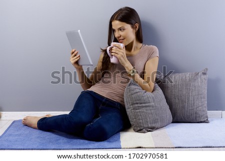 Young woman sitting at home on floor, using tablet computer, drinking tea, smiling.