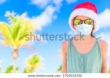 Little happy girl wearing a mask for prevent virus in Christmas hat on white beach