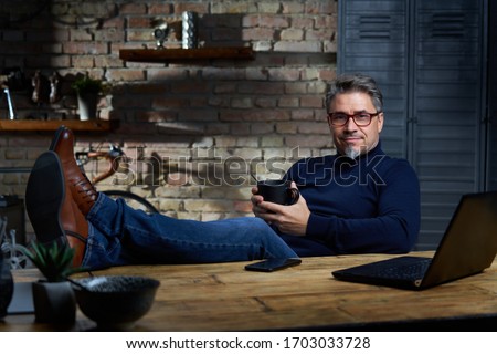 Man sitting at desk at home in easy pose with feet on the table and using phone. Royalty-Free Stock Photo #1703033728