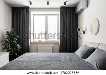 Contemporary bedroom with white walls with a relief picture. There is a double bed with gray linens and pillows, black hanging lamp, window with dark curtains, clock, green plant in the pot, stand.