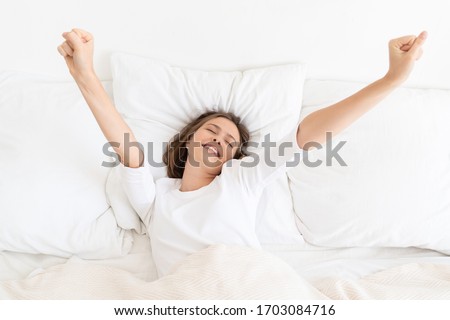 Young woman waking up late in morning on bed with white sheets and pillows, dressed in T-shirt, moving arms trying to get up, eyes closed, smiling Royalty-Free Stock Photo #1703084716
