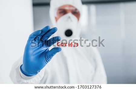 Male doctor scientist in lab coat, defensive eyewear and mask holds glass with covid-19 word on it.
