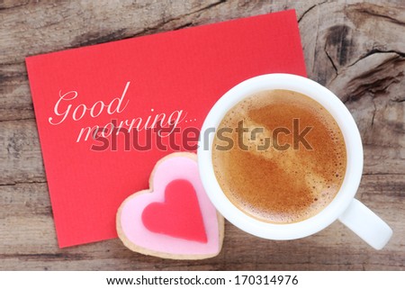 Cup of espresso with creamy foam with a romantic heart shaped cookie and a red greeting card with Good Morning message. Overhead view with copy space