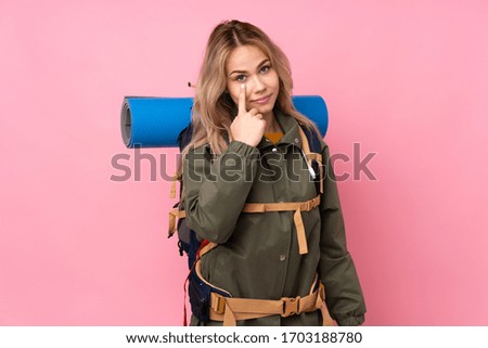 Teenager Russian mountaineer girl with a big backpack isolated on pink background showing something