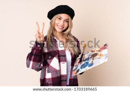 Teenager artist girl holding a palette isolated on blue background showing victory sign with both hands