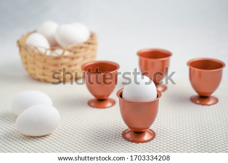 An eggs into a cooper cup and three more empty cooper egg cup behind it, two eggs beside it.,a basket  at bottom