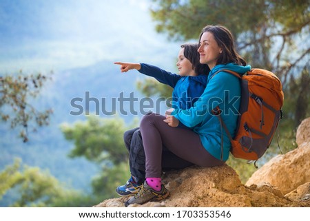 The boy and his mother are sitting on the top of the mountain. A woman is traveling with child. Boy with his mother looking at the mountains. Travel with backpacks. Hike and climb with kids.