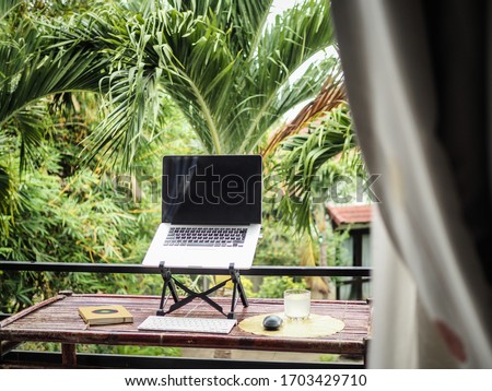 laptop of a remote digital nomad on a wooden bamboo table with notebook, mobile phone and glass in nature on a balcony with a green tropical background with palm trees