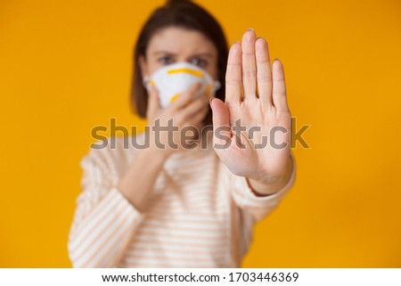 Caucasian woman posing on a yellow wall is gesturing the stop sign while wearing a mask with filter