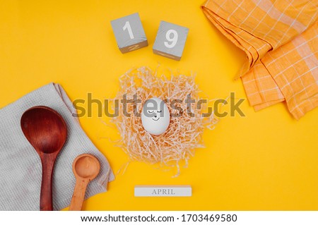 cute Easter egg with a black marker drawn face in a paper nest made of pressed paper, gray wooden cubes with the date of Easter April 19, cotton napkins and wooden spoons on bright yellow background