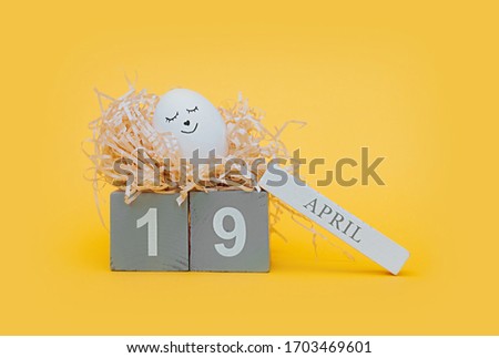cute white Easter egg with a black marker drawn face sleep in a paper nest made of pressed paper on a clear yellow background and gray wooden cubes with the date of Easter April 19