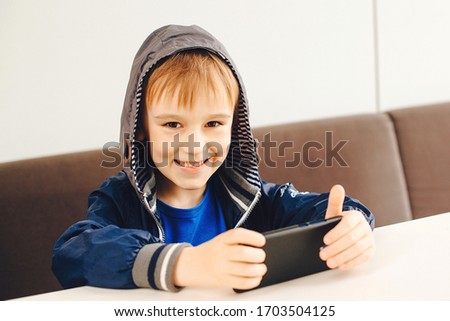 Happy boy playing games on mobile phone. Kid watching videos on smartphone at terrace outside. Modern devices, lifestyle. Stylish schoolboy using smartphone and internet.