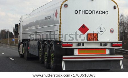 White fuel truck with 33-1203 dangerous class sign and FLAMMABLE inscription in Russian on barrel drive on suburban asphalt highway road at spring day, rear side view - ADR liquid hazardous delivery