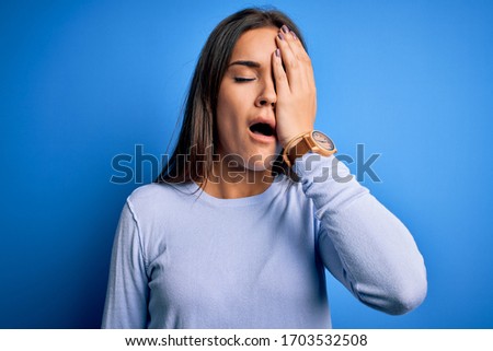 Young beautiful brunette woman wearing casual sweater standing over blue background Yawning tired covering half face, eye and mouth with hand. Face hurts in pain.