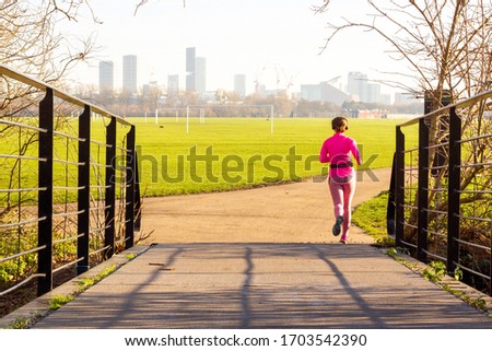 A single woman jogging in pink in Hackney Marshes, with the Olympia Park, London, visible in the distance Royalty-Free Stock Photo #1703542390
