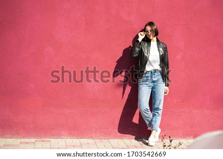 Brunette girl in a sunglasses and a casual clothing (white t shirt, blue jeans and a black jacket), is standing and smiling in front of a pink wall. Picture with a colorful background and a copy space
