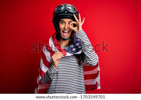 Young beautiful patriotic motorcyclist woman wearing moto helmet and united states flag with happy face smiling doing ok sign with hand on eye looking through fingers