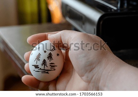 Drawing on a chicken egg. The hand holds a raw egg with a landscape picture on the shell. Close-up. Selective focus.