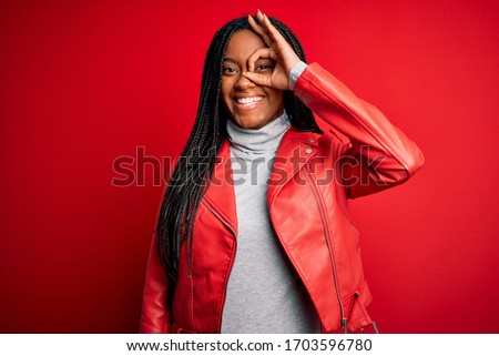 Young african american woman wearing cool fashion leather jacket over red isolated background doing ok gesture with hand smiling, eye looking through fingers with happy face.