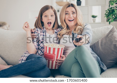 Photo of funny shocked lady mom daughter sit sofa hugging eat popcorn switch on tv set watch movie open mouth intrigue moment stay home quarantine together best friends living room indoor