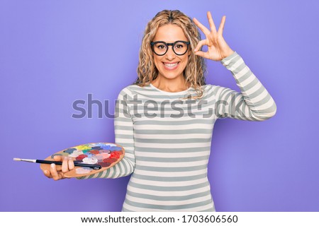 Young beautiful blonde artist woman drawing using paintbrush and palette with colors doing ok sign with fingers, smiling friendly gesturing excellent symbol