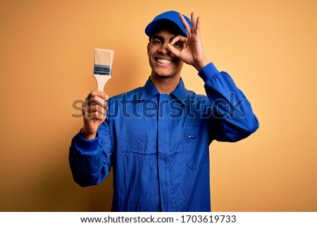 Young handsome african american painter man wearing uniform using painting brush with happy face smiling doing ok sign with hand on eye looking through fingers