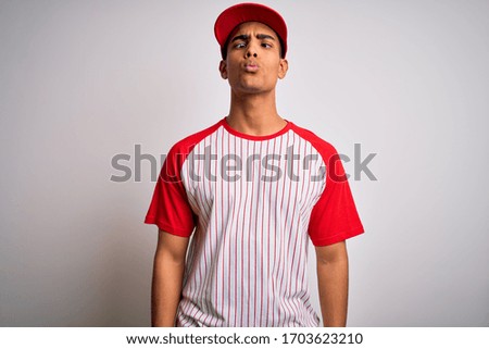Young handsome african american sportsman wearing striped baseball t-shirt and cap making fish face with lips, crazy and comical gesture. Funny expression.