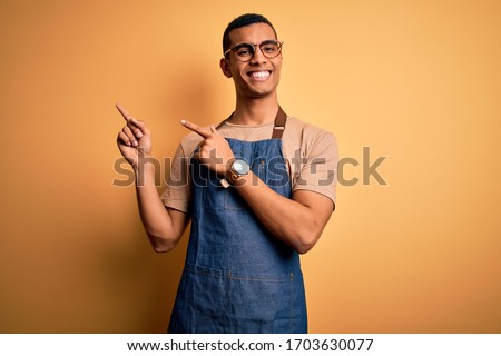 Young handsome african american shopkeeper man wearing apron over yellow background smiling and looking at the camera pointing with two hands and fingers to the side. Royalty-Free Stock Photo #1703630077