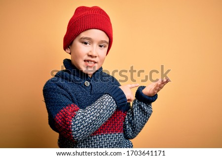 Young little boy kid wearing wool cap and winter sweater over yellow isolated background amazed and smiling to the camera while presenting with hand and pointing with finger.
