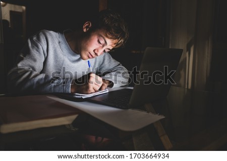 one teenager alone at home doing homework and studying fo an exam in video call with his class and his teacher in quarantine and lockdown - writing with a pencil in a paper with his laptop in front  Royalty-Free Stock Photo #1703664934