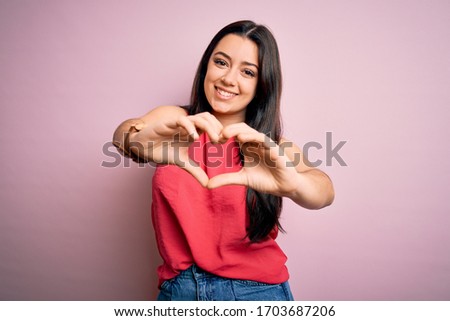 Young brunette woman wearing casual summer shirt over pink isolated background smiling in love doing heart symbol shape with hands. Romantic concept.