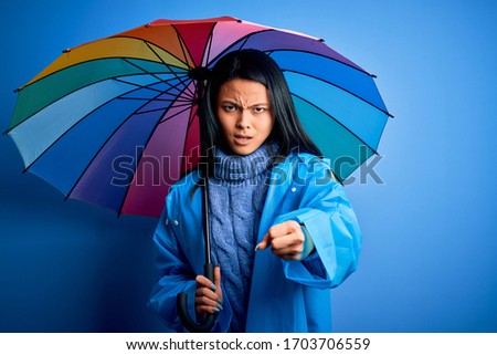 Young chinese woman wearing rain coat holding umbrella over isolated blue background annoyed and frustrated shouting with anger, crazy and yelling with raised hand, anger concept