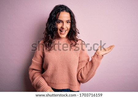 Young beautiful woman with curly hair wearing casual sweater over isolated pink background smiling cheerful presenting and pointing with palm of hand looking at the camera.