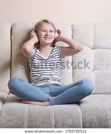A happy teenage girl of 11 years old is sitting on the sofa and listening to music through headphones.  Blonde girl in a striped t-shirt and jeans.
