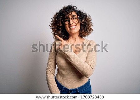 Young beautiful curly arab woman wearing casual t-shirt and glasses over white background cheerful with a smile on face pointing with hand and finger up to the side with happy and natural expression