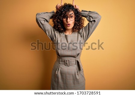 Beautiful arab business woman wearing dress and glasses standing over yellow background doing funny gesture with finger over head as bull horns