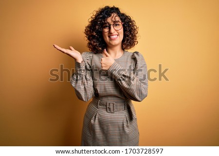 Beautiful arab business woman wearing dress and glasses standing over yellow background Showing palm hand and doing ok gesture with thumbs up, smiling happy and cheerful