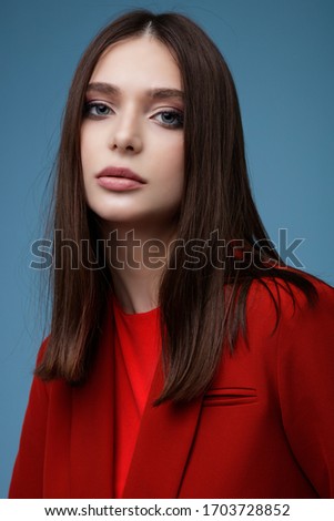 High fashion photo of beautiful elegant young woman in pretty red suit,  jacket, short skirt posing on blue background. Slim figure. Studio shot. 