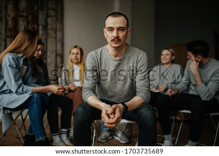 serious caucasian man, member of alcoholics anonymous club sit in center of isolated room. people support each other in the background, sit in the circle. help, support, problem, psychotherapy Royalty-Free Stock Photo #1703735689