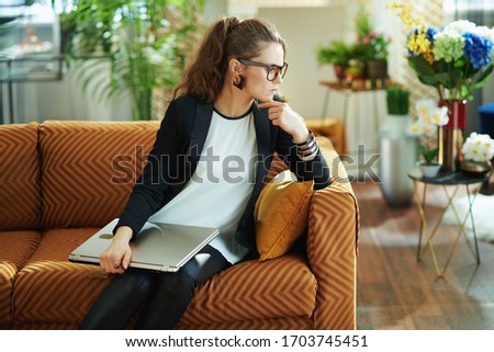 pensive stylish woman in white blouse and black jacket with closed laptop sitting on couch in the modern living room in sunny day.